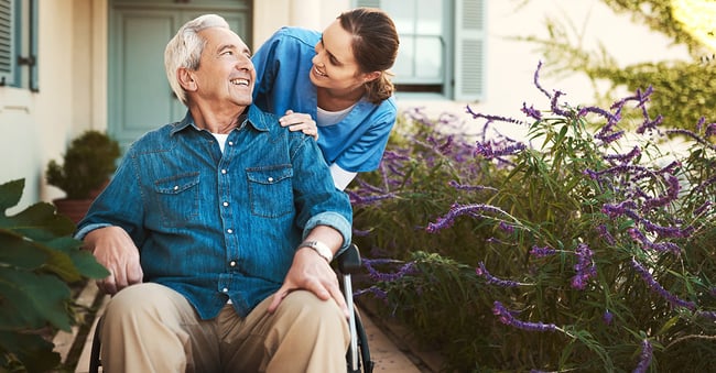 In-home nurse wearing scrubs pushes elderly man on sidewalk outside house