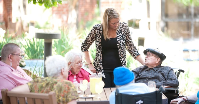 Staff member stands next to seniors in wheelchair at table with friends