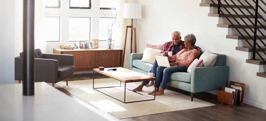 older couple sitting on the couch in their living room looking at laptop and smiling at eachother